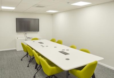 Modern meeting room with a large white table, yellow chairs, and a wall-mounted screen.