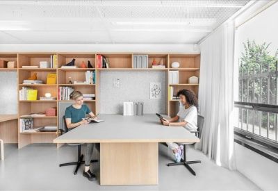 Two people working at a large grey desk in a bright office with built-in wooden shelving.