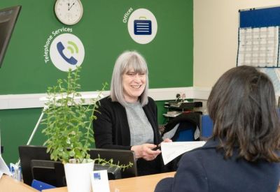 Friendly reception area with a green wall, informative icons, and a receptionist assisting a visitor.