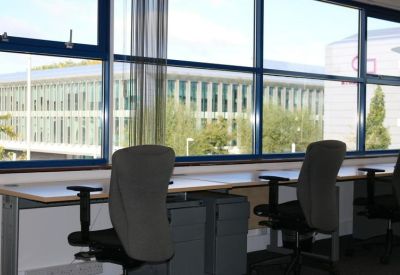 Open-plan office with long desks and grey ergonomic chairs facing large windows.