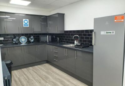 Communal kitchen area with grey cabinetry, black tiled backsplash, and a large silver fridge.