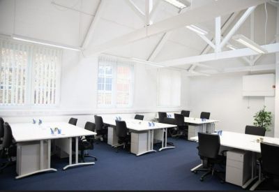 Open-plan office space with multiple white desks, black chairs, and exposed ceiling trusses.