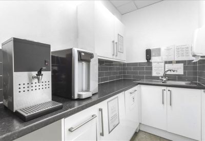 Communal kitchen area with coffee machines, white cabinetry, and grey tiled backsplash.