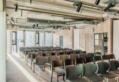 Spacious meeting room with rows of grey chairs, light wood paneled walls, and exposed ceiling ductwork.
