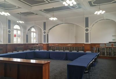 Spacious hall arranged for meetings with a U-shaped table, blue cloths, and tall arched windows.