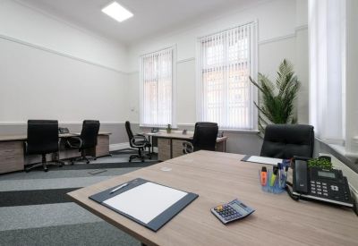 Modern office space with multiple workstations, grey patterned carpet, and tall windows providing natural light.