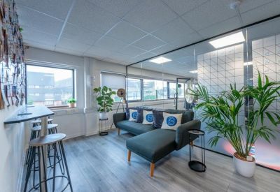 Bright communal lounge area with a grey sectional sofa, potted plants, and natural light.