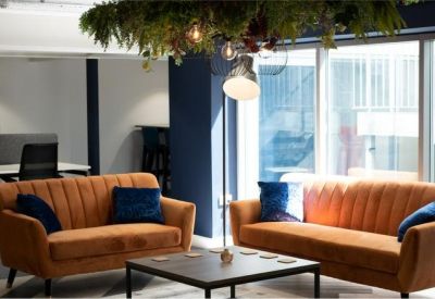 Bright communal lounge with two orange sofas, blue cushions, and a coffee table under hanging plants.