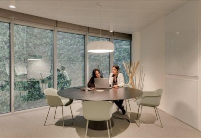 Modern communal area featuring two people working at a round table by large windows.