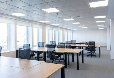 Bright open-plan office with rows of wooden desks and black mesh ergonomic chairs.