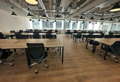 Large open-plan office workspace with rows of wooden desks, black ergonomic chairs, and industrial lighting.
