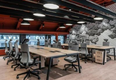 Bright open-plan workspace with multiple wooden desks, grey ergonomic chairs, and red ceiling accents.