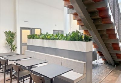 Spacious communal dining area with banquet seating and plants beneath an industrial staircase.
