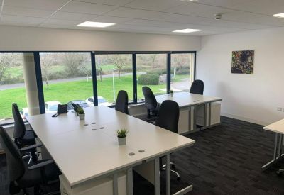 Bright shared office space with white desks, ergonomic black chairs, and large windows overlooking greenery.