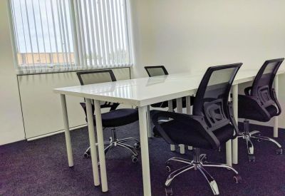Coworking area with white desks and black ergonomic chairs against a window with vertical blinds.
