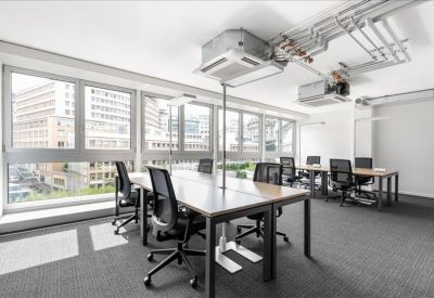 Open-plan office space with large windows and multiple wooden desks with ergonomic chairs.
