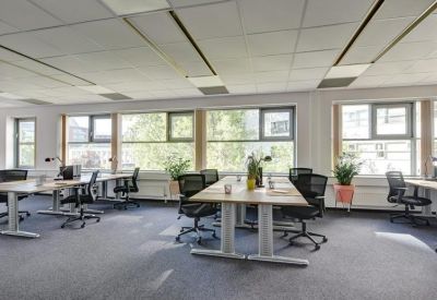 Open-plan office space featuring wooden desks, black ergonomic chairs, and large windows.