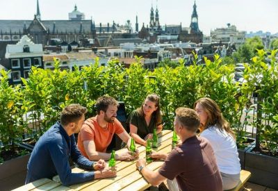 Rooftop terrace with people sitting at a wooden table enjoying drinks with a city skyline view.