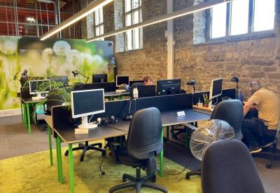 Open-plan office with green-legged desks, black chairs, and exposed brick walls.