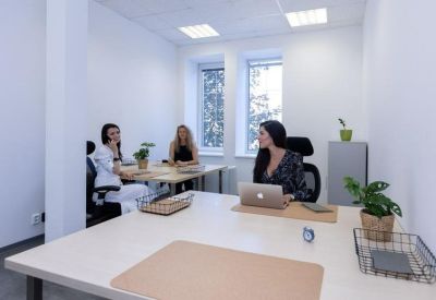 Bright private office suite with three people working at white desks near large windows.