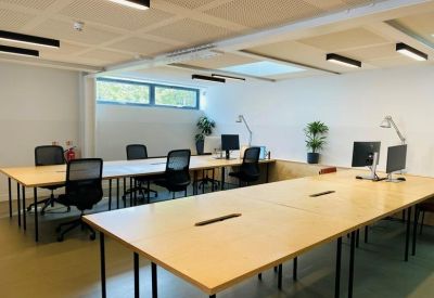 Open-plan office space with long wooden desks, black ergonomic chairs, and natural light.
