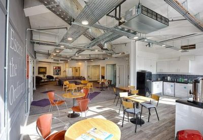 Communal breakroom with tables, chairs, and a kitchen area featuring exposed ceiling ductwork.