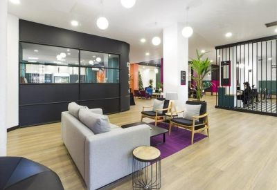 Spacious modern lobby with light wood flooring, a grey sofa, and comfortable lounge chairs.