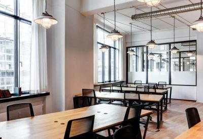 Open-plan office with multiple rows of wooden desks and industrial pendant lighting.