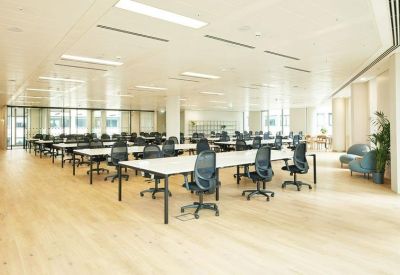 Open-plan office with rows of white desks and black ergonomic chairs on light wood flooring.