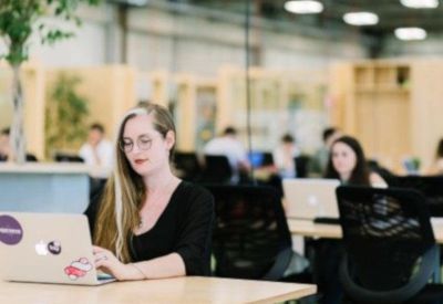 Bright communal lounge area with a woman working on a laptop and plants in the background.