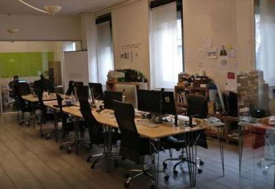 Open-plan workspace with rows of long wooden desks, black ergonomic chairs, and large windows.