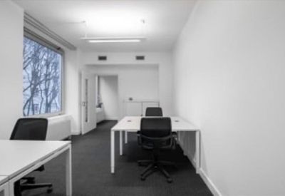 Open-plan office with white desks, black chairs, and views of trees through a large window.