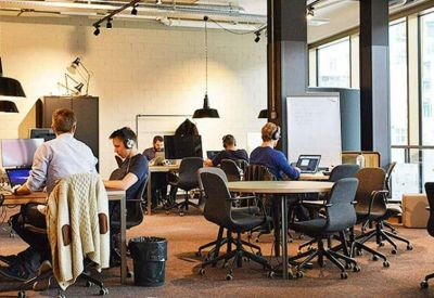 Open-plan office with people working at desks and a round meeting table under industrial lighting.
