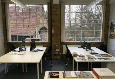 Open-plan office space featuring desks with computer monitors and large industrial-style windows.