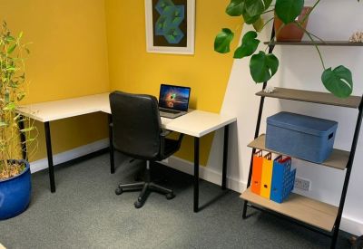 Corner workstation with a white L-shaped desk against a vibrant yellow wall and a plant.