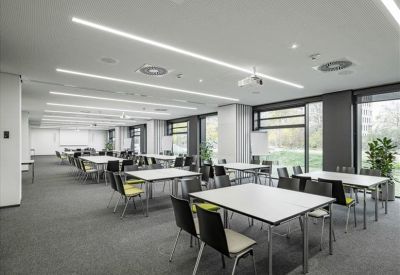 Spacious coworking area with multiple tables, natural light from large windows, and indoor plants.