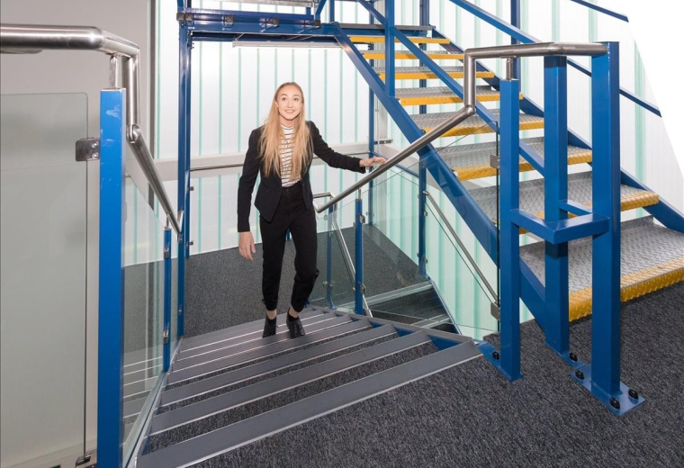 A woman walks down a modern blue industrial staircase with glass balustrades at Bromley Road.
