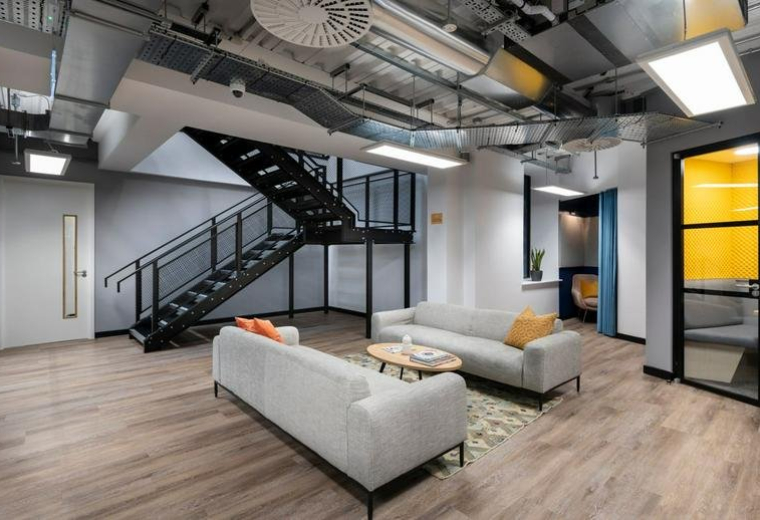 Spacious communal lounge at 33 Foley Street, Fitzrovia featuring grey sofas and a central black staircase.