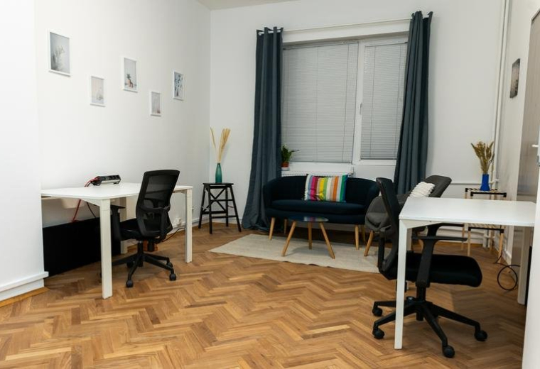Bright office space showing two white desks, ergonomic chairs, and a velvet sofa near the window.