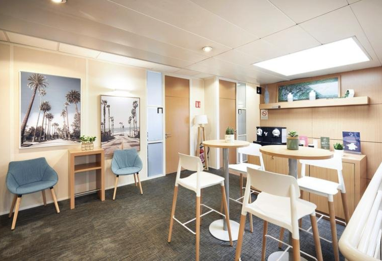 Communal breakout area at Europarc de Pichaury with high-top tables, blue armchairs, and tropical wall art.