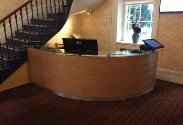 Curved wooden reception desk with a computer monitor located near a classic staircase.