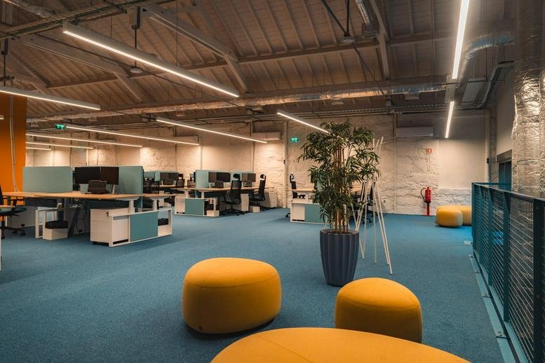 Open-plan workspace at 584, Rua Brito e Cunha featuring industrial ceilings, blue carpets, and yellow seating.