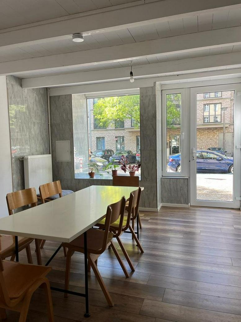 Communal dining area with a long white table, wooden chairs, and views of the street through a large window.