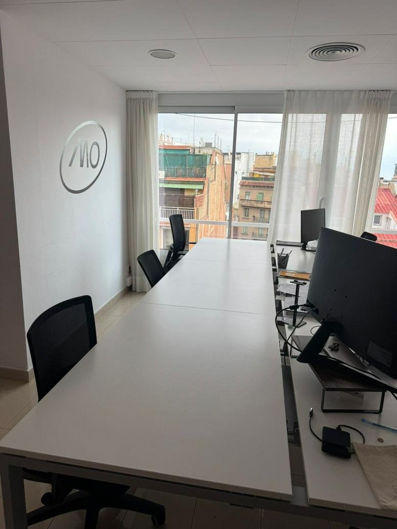 Long white shared workspace desks with black chairs and city views at 374, Carrer de Muntaner, Barcelona.