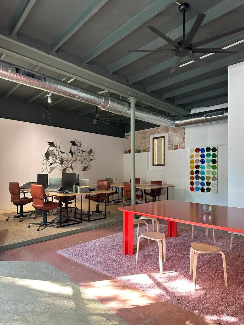 Sunlit communal area featuring a long red table and modern wooden stools.