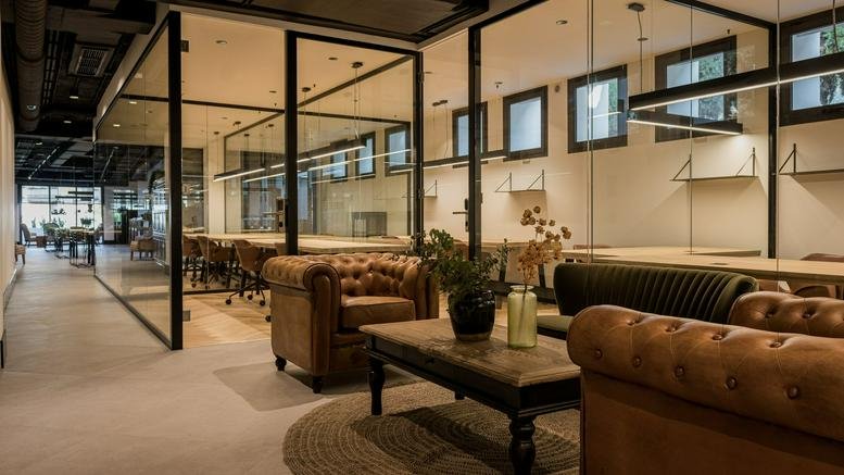 Bright glass-walled workspace featuring leather chesterfield sofas and long wooden desks.