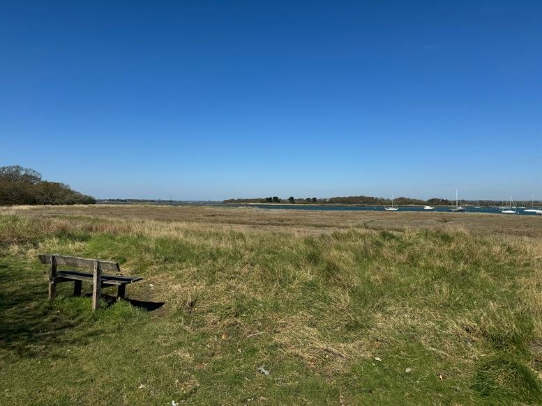 Coastal view from Northshore, Itchenor, Chichester, England featuring a wooden bench and grassy marshland.