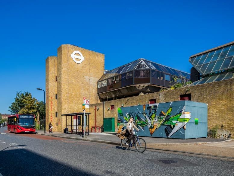 Modern exterior of Hackney Depot at 5 Sheep Lane with unique glass architecture and a red bus passing.