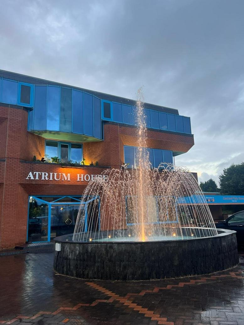 Wide exterior shot of Atrium House at dusk with a central illuminated fountain and blue glass facade.
