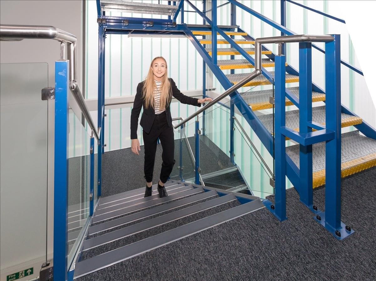 A woman walks down a modern blue industrial staircase with glass balustrades at Bromley Road.